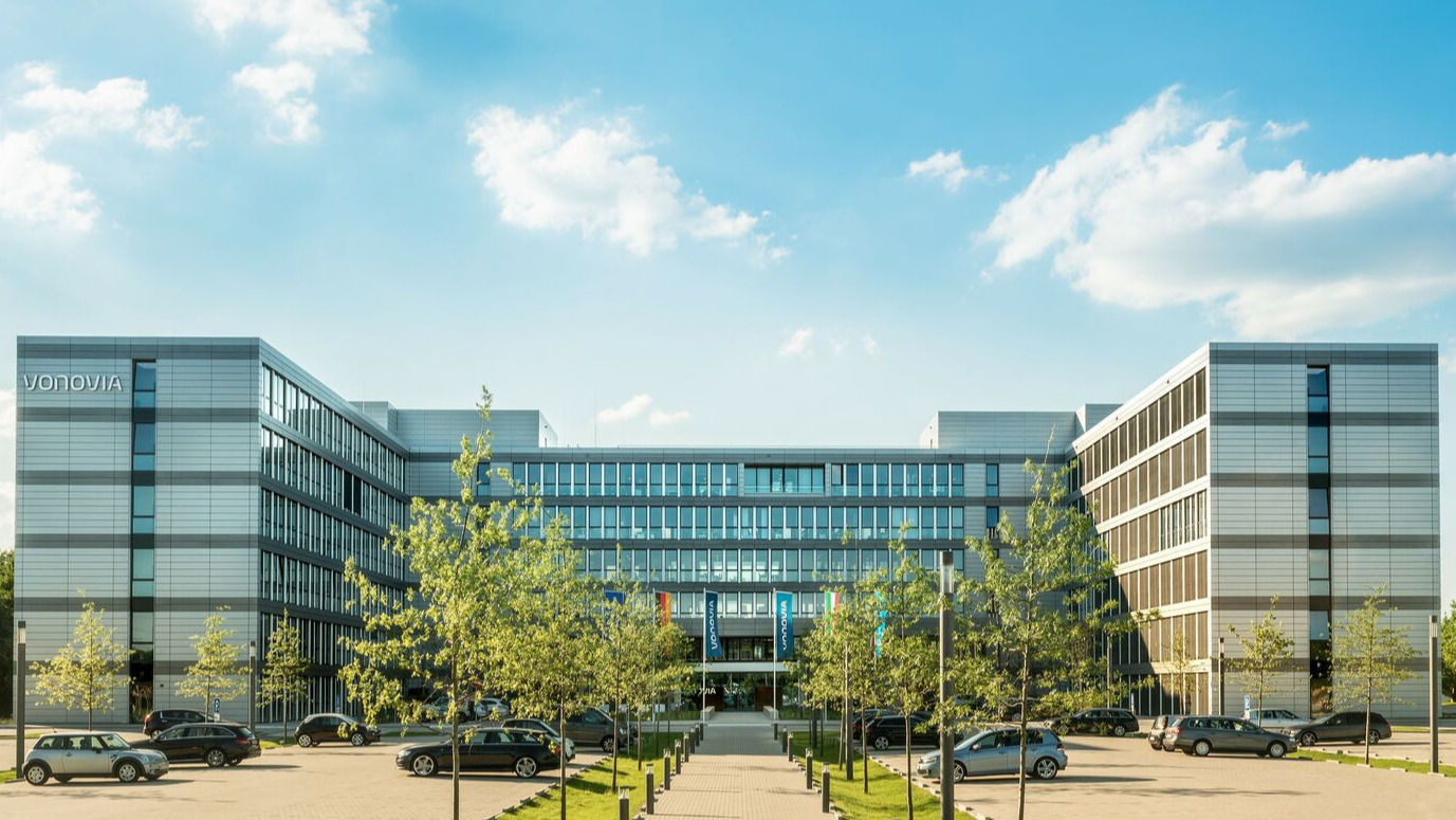 Großes, modernes Gebäude mit Vonovia Schriftzug, Autos im Vordergrund, leicht bewölkter Himmel